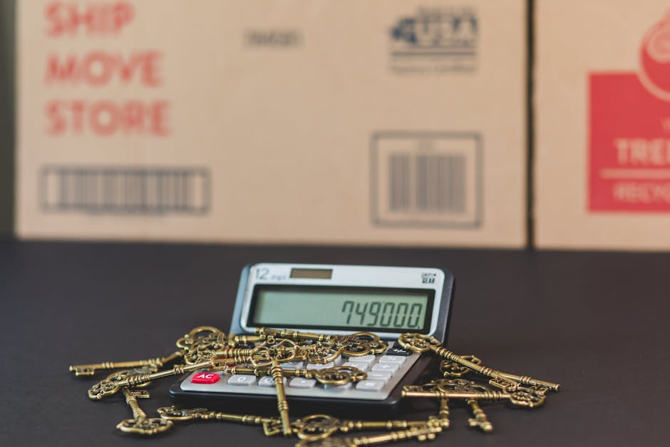 A close-up view of a white calculator with a large digital display showing the number 749800, placed on a dark surface in front of stacked cardboard moving boxes marked with red and black text, including 'Ship', 'Move', and 'Store'. Several antique brass keys and keychains are scattered around the calculator, some partially overlapping it, indicating a packing or property relocking process associated with house removals. In the background, the boxes are positioned behind the calculator and keys, suggesting a home relocation environment. This setup visually reflects the logistical aspects of furniture transport and moving, with a focus on packing materials and secure item handling, aligned with professional removal services provided by Man and Van Lambeth.