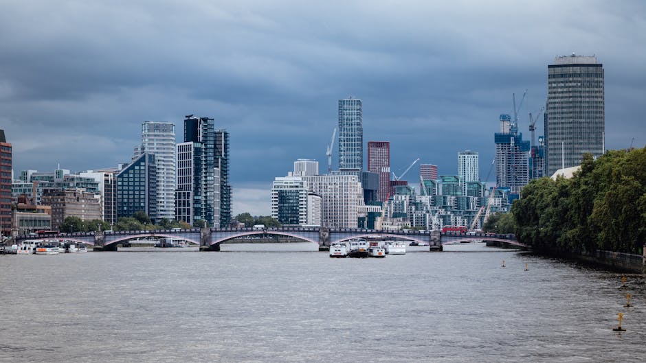 A cityscape view of a river with several boats docked along the shoreline, leading towards a dense cluster of modern skyscrapers and office buildings under a cloudy sky, with some cranes visible indicating ongoing construction. To the right, a green tree-lined area borders the water, creating a contrast with the urban environment. The scene captures the environment surrounding a home relocation or moving operation, where equipment such as trolleys or packing materials might typically be used during furniture transport and packing processes. The image is relevant to house removals services in Lambeth, reflecting the infrastructure and urban setting involved in the logistics of moving, and is hosted on a website focusing on transparent, no hidden fees removals in the area.