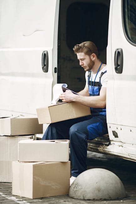 A young man wearing a striped t-shirt and blue overalls is seated at the open side door of a white moving van, surrounded by cardboard boxes of various sizes, some stacked on the ground and others inside the van. He is holding a document or checklist and appears to be reviewing packing or loading details. The scene depicts the preparation phase of a house removal, with the van parked on a paved area. The background includes a concrete bollard and the interior of the van, which is ready for furniture transport, highlighting the logistics involved in packing and moving. Man and Van Lambeth provides professional removals services, supporting efficient home relocation and furniture transport through careful planning and organization.