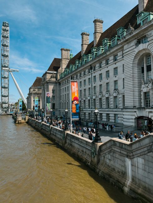 A daytime view of a historic riverside building with a traditional architectural style, featuring multiple chimneys, large windows, and decorative stonework, situated along the Thames River in Lambeth. The building's white facade contrasts with its dark roof and green window frames. Along the riverbank, a crowded walkway is lined with lampposts and bordered by a low stone wall, with pedestrians walking and gathering. A row of large balloons and banners displays advertisements, including a notable character resembling Mario from a popular video game, which are attached to tall, slim poles extending above the walkway. A modern Ferris wheel is visible in the background on the left, indicating an urban, lively environment. The scene captures a clear, sunny day with a bright blue sky, enhancing the visibility of various architectural and crowd details, relevant to the context of Lambeth's urban setting and potential home relocation logistics from nearby residential and commercial areas, where Man and Van Lambeth offers removal and moving services.