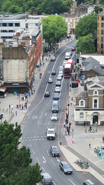 An aerial view of a tree-lined urban street with parked cars along the curb and moving vehicles, including a double-decker bus, on the road. The street is adjacent to a mix of older and modern buildings, some with brick facades and others with flat roofs, with pedestrians walking on the sidewalks. At the corner, there are bicycles parked near a small building, and a few people are visible walking or waiting at crosswalks. The scene is well-lit with natural daylight, capturing the busy atmosphere typical of a central residential area in Lambeth, suitable for house removals or home relocation services provided by Man and Van Lambeth.