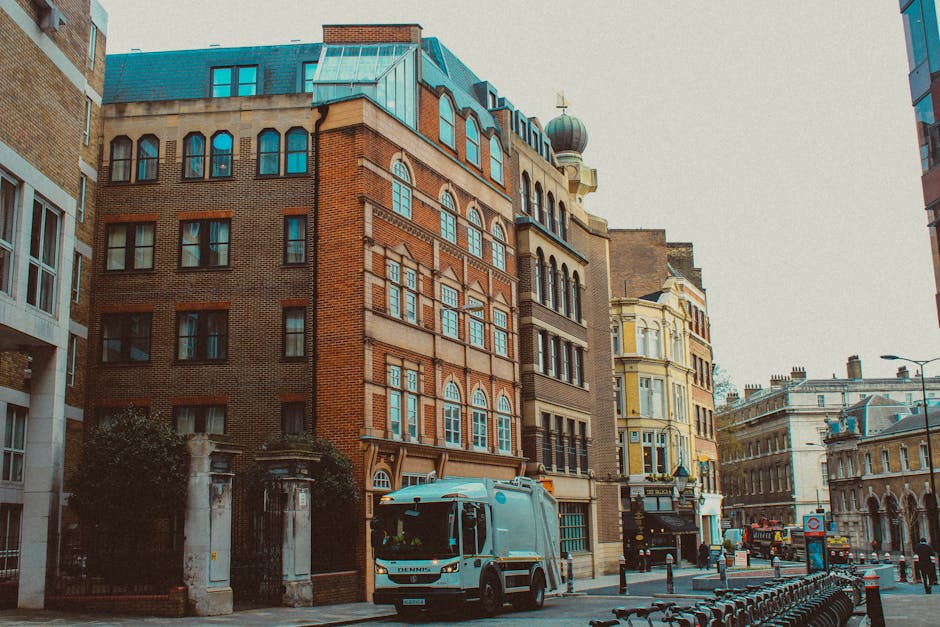 A street scene outside a historic multi-storey building with red brick and ornate architectural details, situated in an urban area with other similar buildings. In the foreground, a light blue van belonging to Man and Van Lambeth, a professional removals company, is parked near a curb, positioned for loading or unloading. Behind the van, multiple large cardboard boxes and wrapped furniture are arranged on the pavement, indicating preparations for a house relocation. The boxes are sealed with packing tape and some are covered with protective plastic or fabric wraps. Nearby, a worker is using a hand truck or trolley to assist in transporting boxes and furniture, with the loading process underway. The scene is lit with natural daylight, capturing the busy logistics involved in furniture transport and moving services in central London, with the environment featuring street lamps, bike racks, and other urban infrastructure.