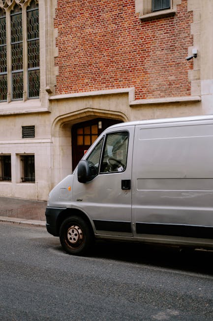 A silver cargo van parked on a residential street in front of a brick building with large, historic windows and an arched stone doorway. The van is positioned close to the curb, with the driver's side facing the building. No other vehicles or pedestrians are visible. The scene suggests a home relocation process, with the van potentially being used by Man and Van Lambeth for furniture transport and packing and moving services. The environment is in daylight, with natural lighting illuminating the urban setting, and the van appears ready for loading or unloading items such as boxes, furniture, or appliances, supporting efficient removals and relocation work.