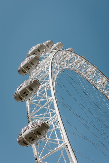 A large white observation wheel, with multiple enclosed cabins made of transparent material and white framing, is seen from a low-angle perspective against a clear blue sky. The structure features an intricate network of white metal supports and spokes, supporting the cabins along the outer edge of the wheel. The cabins are evenly spaced, some of them visible as they are attached to the wheel's circumference. The photograph captures the upper part of the wheel, focusing on the curvature and the engineering details of the support framework, highlighting the height and scale of the structure typically associated with city attractions or landmarks used in urban leisure activities. This image provides a clear view of the wheel's external design and its prominent position in the skyline, relevant to the concept of transport, sightseeing, or large-scale attractions involved in moving or setting up such equipment as part of home or commercial relocation or event logistics, as seen in professional removal services like those offered by Man and Van Lambeth.