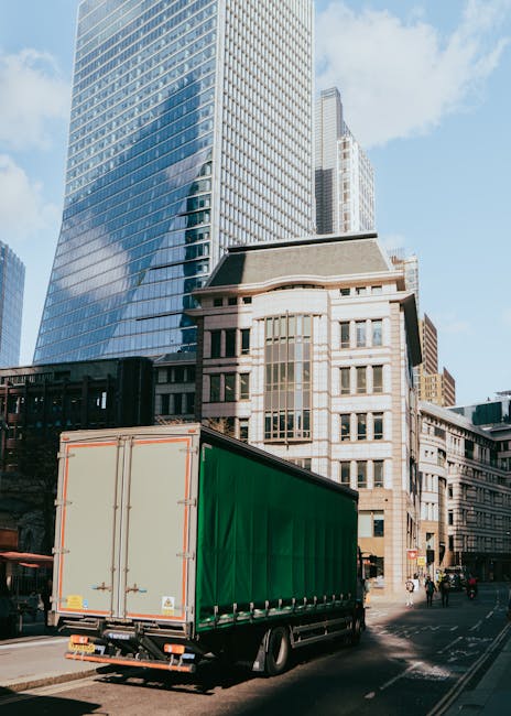 A large green-draped moving truck parked on a city street in front of tall modern office buildings with glass facades and a historic stone building with multiple windows. The truck is positioned close to the curb, with the rear doors closed, indicating it is either preparing for or in the process of a house or office removal. The surroundings include a paved roadway, pedestrians, and a clear sky, suggesting daytime. This scene illustrates the logistics involved in furniture transport and packing during a professional relocation, with [COMPANY_NAME] providing moving services in the Lambeth area as seen on the webpage about office removals from Waterloo to Southbank.
