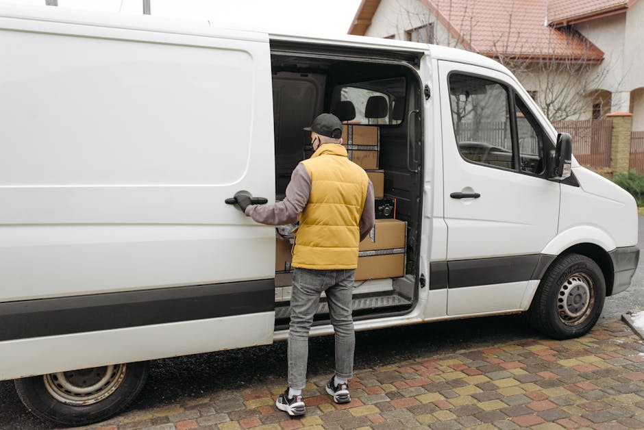A man dressed in a yellow puffer vest, grey long-sleeve shirt, grey jeans, and black sneakers with white soles is standing next to a white commercial van, holding the sliding side door open. Inside the van, several cardboard boxes of various sizes are neatly stacked, some with black straps securing them. The boxes are brown, with some wrapped in clear plastic or covered with packing paper inside the vehicle. The van is parked on a brick-paved driveway of residential houses with wooden fences and a small tree visible in the background. The scene captures a home relocation process step, with the man preparing to load or unload furniture and packed items, supported by the professional removals service provided by Man and Van Lambeth, which specialises in home removals and furniture transport in the Lambeth and Vauxhall areas.