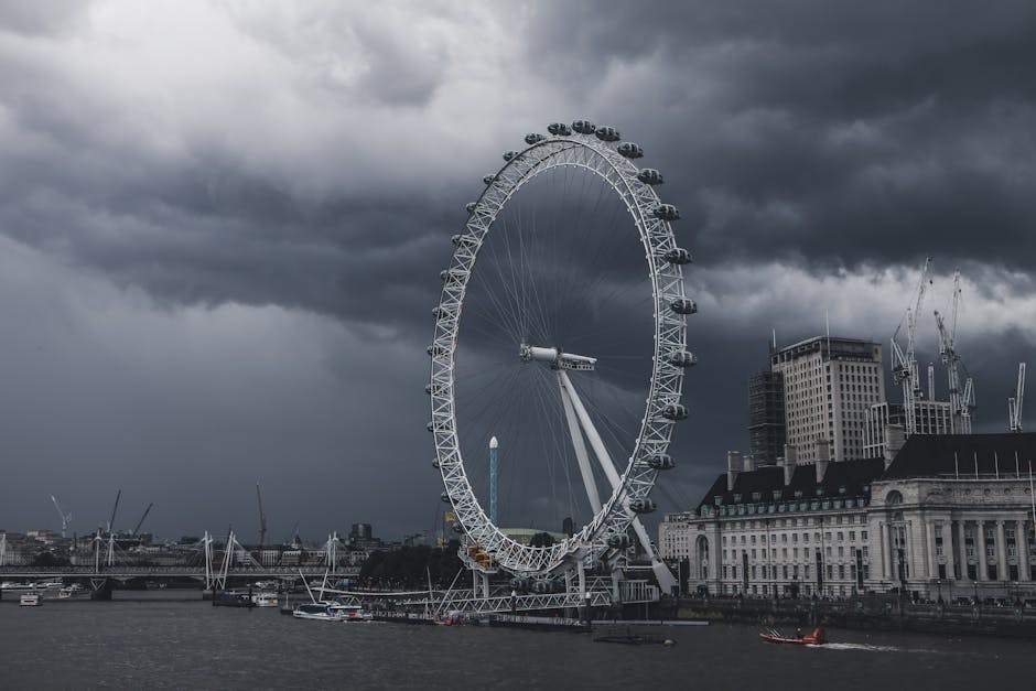 A photograph of the London Eye, a large Ferris wheel, situated on the south bank of the River Thames in London. The wheel features multiple enclosed passenger capsules attached around its circumference, visible against a backdrop of dark, stormy clouds. In the foreground, the riverbank includes boats docked along the water, with some partially obscured. To the right of the London Eye, several cranes and high-rise buildings under construction are visible, indicating ongoing urban development. The scene is illuminated by natural, subdued lighting due to the cloudy sky, adding a dramatic atmosphere. The image captures the iconic promenade area where the London Eye is located, aligning with themes of city attractions and transportation. This visual is relevant for services related to urban relocation, packing and moving logistics, or city-based removals, with the presence of the Ferris wheel emphasizing infrastructure and transport-related visuals. Man and Van Lambeth, known for removals services, may use such imagery to convey transportation and moving processes within an urban context.