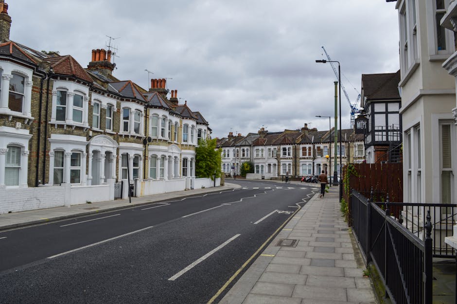 An empty residential street in a suburban area with rows of Victorian-style terraced houses on the left, featuring white-painted facades, bay windows, and red brick chimneys. The pavement on the right has a black metal fence, with a white building partially visible. The street is paved with dark asphalt, marked with white roadside lines, and has typical London-style street lighting and a construction crane visible in the background. The sky is overcast with gray clouds, creating a subdued lighting environment. This scene depicts a quiet urban environment suitable for house removals and furniture transport, with no visible moving activity but providing context for vehicle access and parking associated with home relocation services by Man and Van Lambeth.