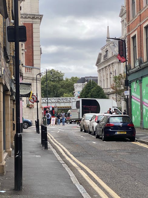 A narrow urban street in Lambeth with a row of parked cars along the right side, including a black vehicle closest to the camera. The sidewalk on the left is lined with black metal posts and features small businesses, one with a sign displaying 'Emilia.' In the background, a white moving van is parked near the curb with its rear doors open, partially blocking the view of the street beyond. Several people are visible near the van, appearing to be engaged in loading or unloading furniture and boxes wrapped in plastic and cardboard. The area is shaded by overcast skies, and taller historic buildings, including a church with a steeple and a classical-style structure, rise above the street scene, indicating a London neighbourhood scene involved in home relocation or furniture transport services such as those offered by Man and Van Lambeth.
