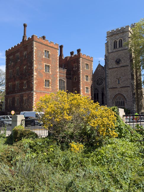 A historic brick building with multiple rectangular windows, topped with chimneys and decorative battlements, situated behind a garden area with yellow-flowered shrubs and green foliage. To the right of the building, there is a stone church with a tall, square clock tower featuring arched windows and a crenellated parapet. The scene is set outdoors in bright daylight with a clear blue sky, and a metal fence borders the garden area. In the foreground, several parked vehicles are visible along the street, and the overall scene depicts an urban environment during a home relocation or furniture transport process, typical of house removals or moving services handled by companies such as Man and Van Lambeth, as referenced on the webpage about urgent same day removals in Lambeth.
